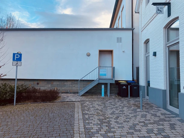IMG_7512.jpeg Ein barrierefreier Eingang mit Rampe und Parkplätzen, zugänglich für Rollstuhlfahrer, an einem Gebäude.
