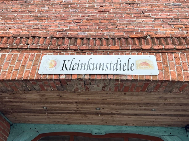 Das Bild zeigt das Schild der Kleinkunstdiele, aufgehängt an einer Backsteinwand mit Holzbalken.