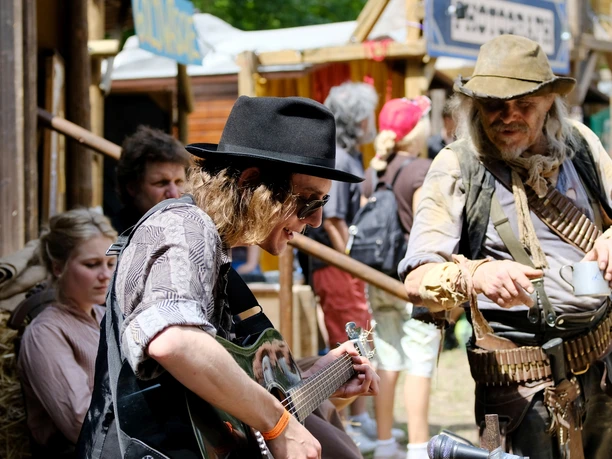Westernstadt3.jpg Musiker spielt Gitarre in authentischer Westernkleidung, umgeben von Zuschauern in Westernkulisse.