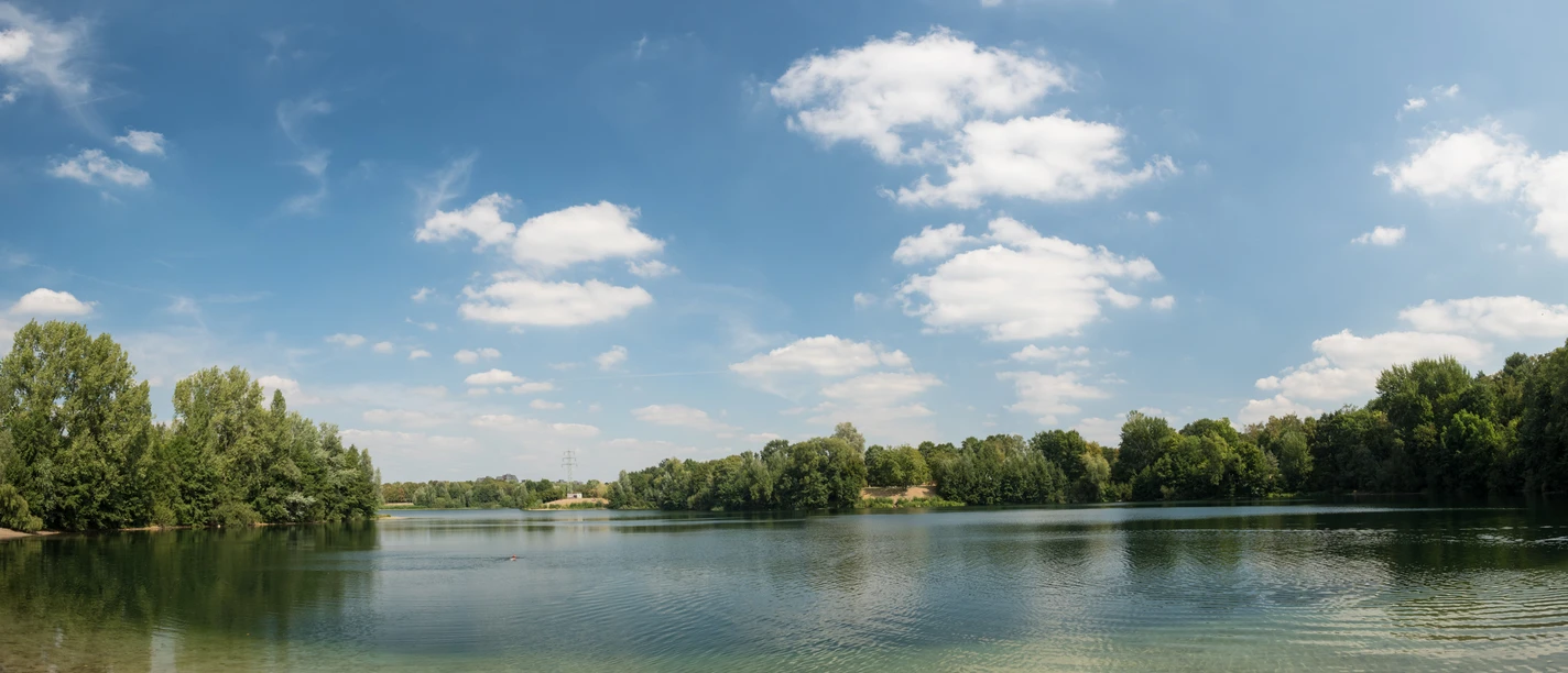 Grüner See im Erholungspark Volkardey in Ratingen Ein klarer See umgeben von dicht bewaldeten Ufern, mit einem strahlend blauen Himmel darüber.