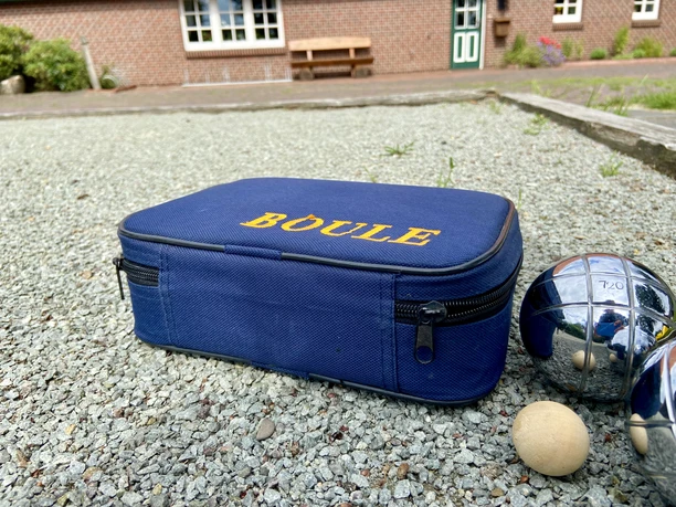 image00004.jpeg Eine Boule-Tasche aus blauem Stoff mit der Aufschrift "BOULE" und drei glänzenden Metallkugeln auf Kies.