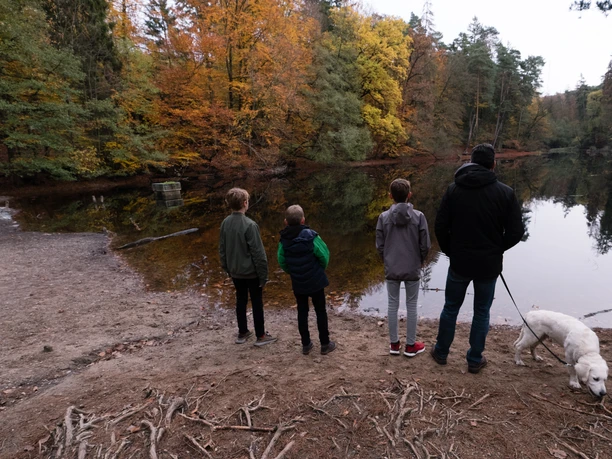 Vier Personen und ein Hund stehen am Ufer eines Sees im Herbst, umgeben von buntem Laub.