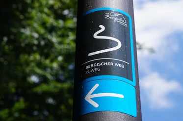 Zuweg Bergischer Weg Das Bild zeigt ein Schild des "Bergischer Weg" Zuwegs vor unscharfem grünem Hintergrund und blauem Himmel.