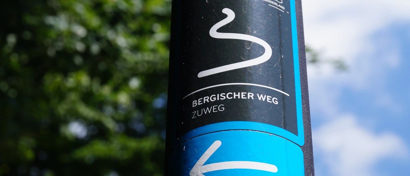 Zuweg Bergischer Weg Das Bild zeigt ein Schild des "Bergischer Weg" Zuwegs vor unscharfem grünem Hintergrund und blauem Himmel.
