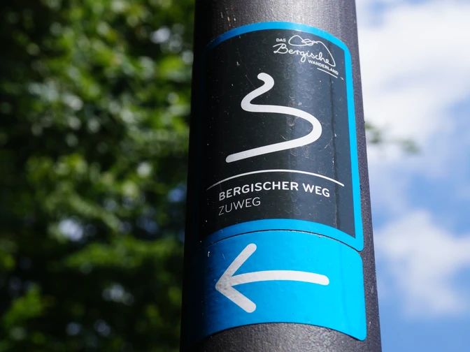 Zuweg Bergischer Weg Das Bild zeigt ein Schild des "Bergischer Weg" Zuwegs vor unscharfem grünem Hintergrund und blauem Himmel.