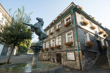 Pferdebrunnen und Stadtgeschichtshaus in Mettmann Bronzefarbene Pferdeskulptur erhebt sich über einem Wasserbrunnen vor einem Fachwerkhaus mit Blumenschmuck.