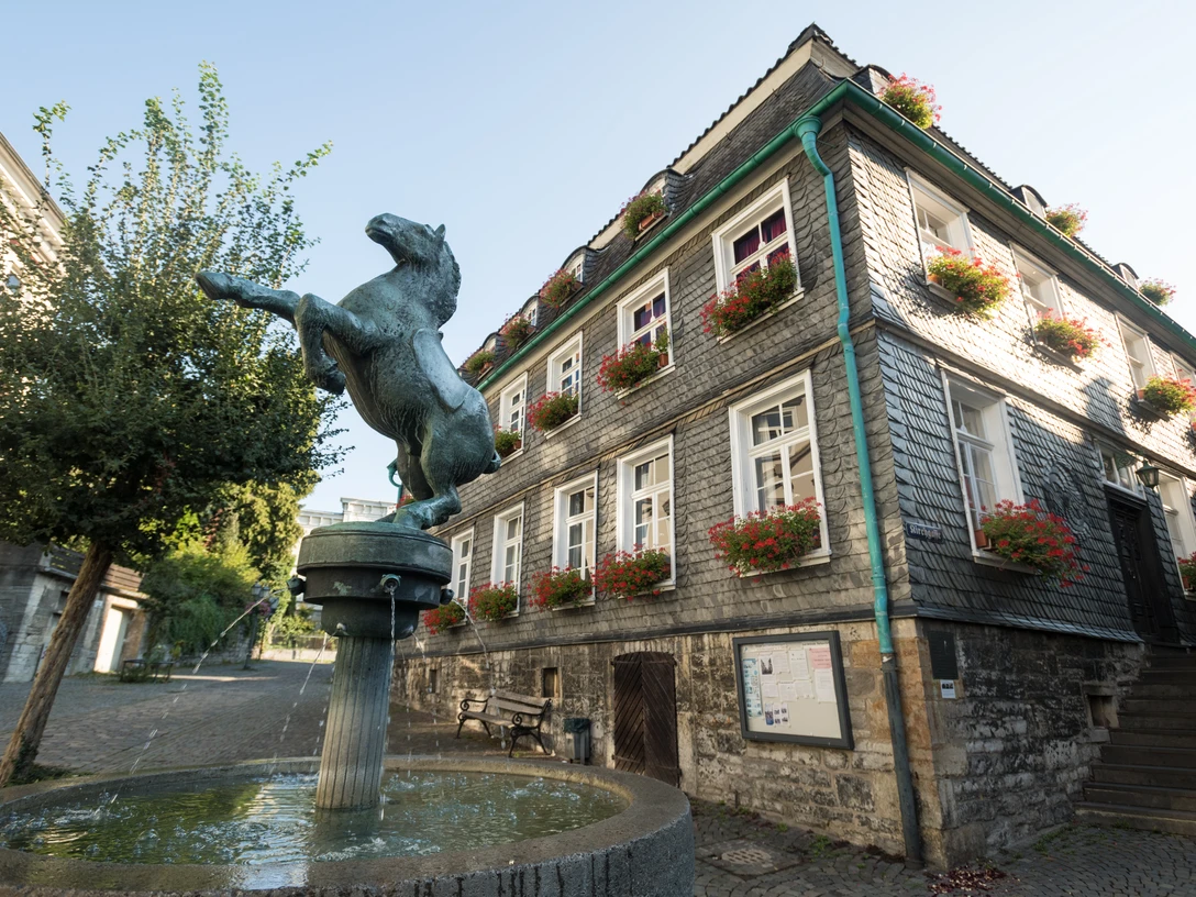 Pferdebrunnen und Stadtgeschichtshaus in Mettmann Bronzefarbene Pferdeskulptur erhebt sich über einem Wasserbrunnen vor einem Fachwerkhaus mit Blumenschmuck.