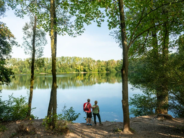 Blick auf den Menzelsee bei Hilden Zwei Personen stehen am Waldufer und blicken auf den ruhigen Menzelsee mit grüner Vegetation.