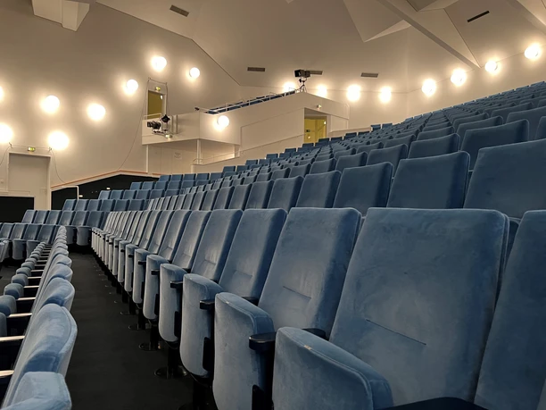 Theater auf dem Hornwerk Reihen aus blauen Theatersitzen blicken auf eine Bühne unter warmem Licht in einem modernen Raum.