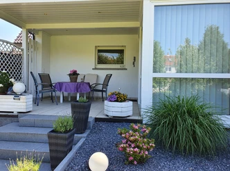 Sonnige Terrasse mit Tisch und Stühlen, umgeben von blühenden Pflanzen und gepflegtem Garten.