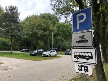 Wohnmobilstellplatz Bücken Ein Parkplatz mit Schildern für Autos, Busse und Wohnmobile, umgeben von Bäumen und geparkten Fahrzeugen.