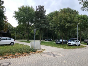 Wohnmobilstellplatz umgeben von Bäumen, mit geparkten Autos und gepflasterten Wegen im Grünen.