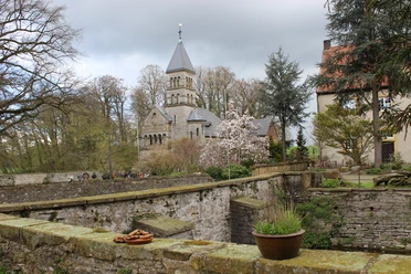 Schloss Brincke.jpg Historische Kirche mit spitzem Turm, blühende Bäume und alte Mauer in malerischer Frühlingslandschaft.