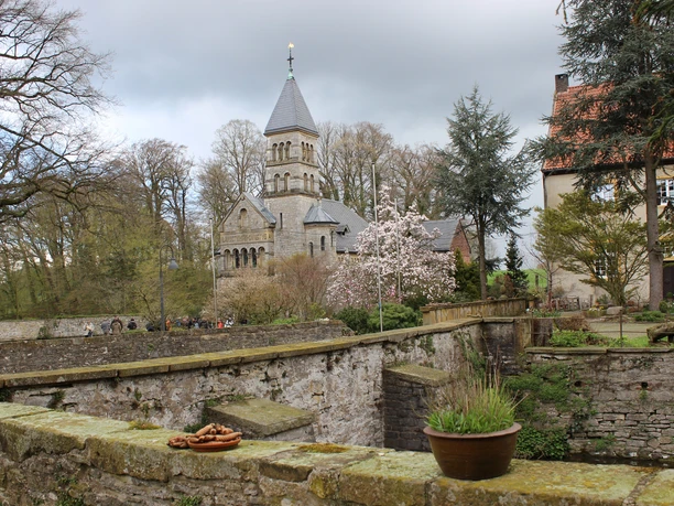 Schloss Brincke.jpg Historische Kirche mit spitzem Turm, blühende Bäume und alte Mauer in malerischer Frühlingslandschaft.