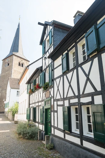Abstecher in die Hildener Innenstadt Fachwerkhäuser und ein Kirchturm entlang einer gepflasterten, grünen Gasse in der Hildener Innenstadt.