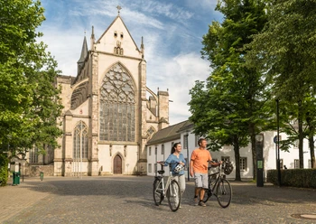 Rad fahren in Altenberg <p>Ein Paar schiebt Fahrräder vor dem Altenberger Dom bei sonnigem Wetter, umgeben von Bäumen.</p>