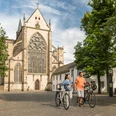Rad fahren in Altenberg <p>Ein Paar schiebt Fahrräder vor dem Altenberger Dom bei sonnigem Wetter, umgeben von Bäumen.</p>