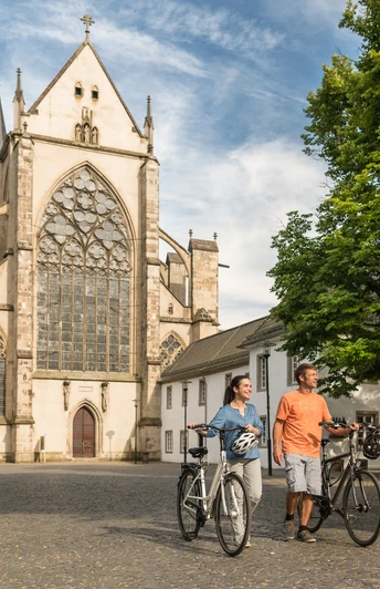 Rad fahren in Altenberg <p>Ein Paar schiebt Fahrräder vor dem Altenberger Dom bei sonnigem Wetter, umgeben von Bäumen.</p>