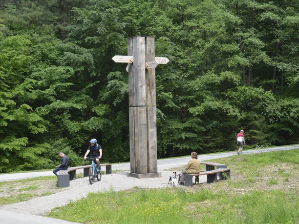 Ein hölzernes Wanderkreuz mit Wegweisern steht vor grünen Bäumen, umgeben von Radfahrern und Sitzbänken.