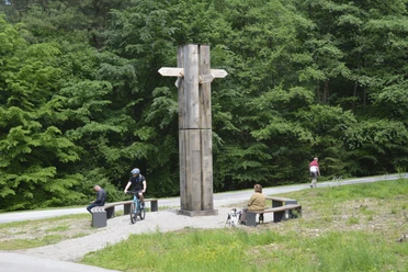 Wanderkreuz (3) @LTM GmbH.JPG Ein hölzernes Wanderkreuz mit Wegweisern steht vor grünen Bäumen, umgeben von Radfahrern und Sitzbänken.