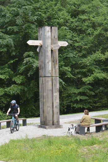 Wanderkreuz (3) @LTM GmbH.JPG Ein hölzernes Wanderkreuz mit Wegweisern steht vor grünen Bäumen, umgeben von Radfahrern und Sitzbänken.