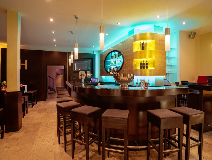 Bar.jpg Moderne Hotelbar mit stimmungsvollem Licht, dunklen Holzverkleidungen und gepolsterten Barhockern.
