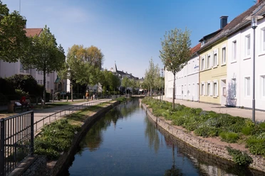 Parklandschaft Friedrichstal Kanal in Friedrichstal, gesäumt von Bäumen und Häusern, unter blauem Himmel an einem sonnigen Tag.