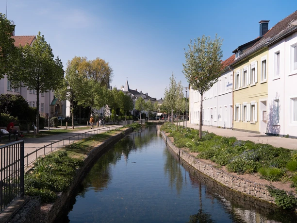 Parklandschaft Friedrichstal Kanal in Friedrichstal, gesäumt von Bäumen und Häusern, unter blauem Himmel an einem sonnigen Tag.