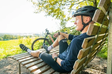 Mountainbike Eine Person entspannt auf einer Holzliege neben einem Fahrrad, mit Blick auf eine grüne Landschaft.