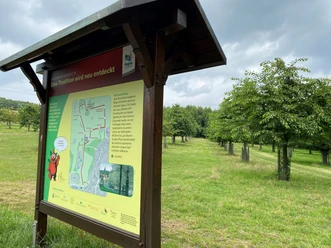 Lehrpfadschild.jpg Informatiebord met wandelkaart in een groen weidelandschap onder een licht bewolkte hemel.