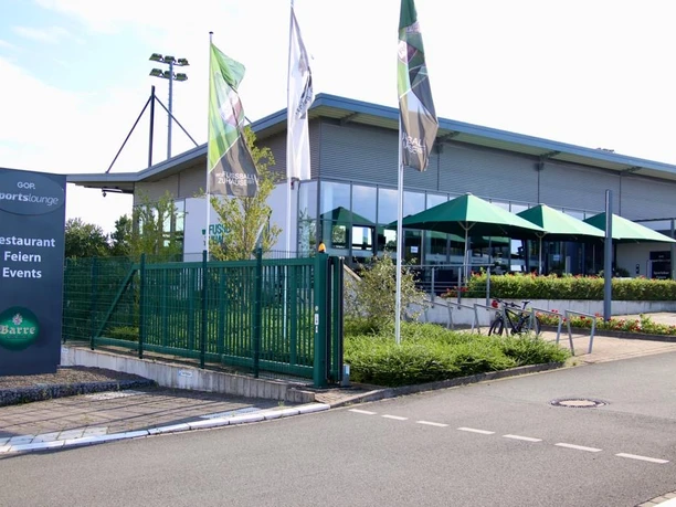 GOP Sportslounge mit moderner Glasfassade, grünen Sonnenschirmen und umgebenden Werbefahnen.