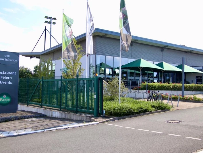 Außenansicht GOP Sportslounge GOP Sportslounge mit moderner Glasfassade, grünen Sonnenschirmen und umgebenden Werbefahnen.