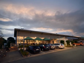 Außenansicht GOP Sportslounge Moderne Sportslounge mit großer Glasfassade und markanten grünen Sonnenschirmen im Eingangsbereich.