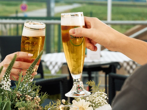 Getränke Terrasse Zwei Personen stoßen mit Biergläsern an einem weiß gedeckten Tisch im Freien vor landschaftlichem Hintergrund an.