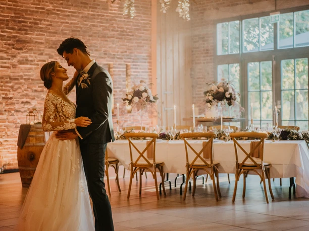 Hochzeit Ein Brautpaar tanzt in einem elegant dekorierten Raum mit festlich gedecktem Tisch im Hintergrund.