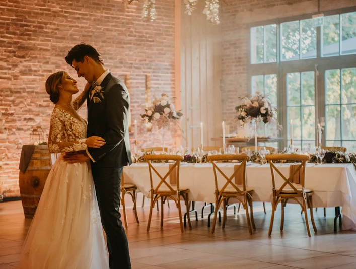 Hochzeit Ein Brautpaar tanzt in einem elegant dekorierten Raum mit festlich gedecktem Tisch im Hintergrund.