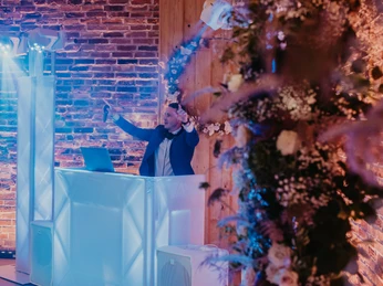 Scheune - Eventlocation DJ in Anzug steht hinter beleuchtetem Pult, umgeben von Backsteinwänden und dekorativen Blumen.
