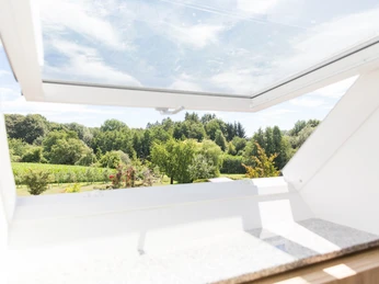 Comfort-Zimmer Ausblick ins Grüne durch ein geöffnetes Dachfenster, sonniger Himmel, umgeben von Bäumen und Wiesen.