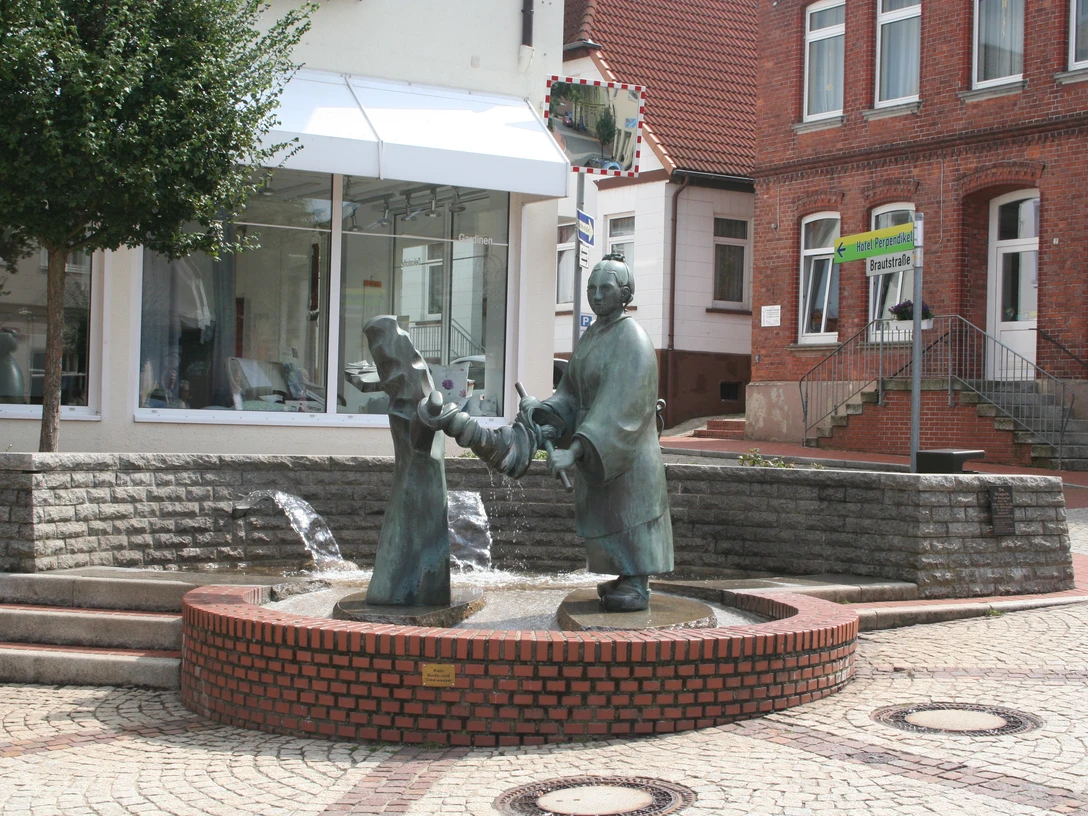 Bronzefigur einer Frau beim Auswringen von Wäsche auf einem Brunnen in einem gepflasterten Stadtplatz.