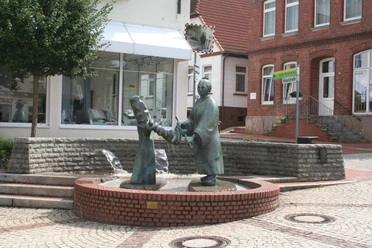 Brunnen - Die Wringerin Bronzefigur einer Frau beim Auswringen von Wäsche auf einem Brunnen in einem gepflasterten Stadtplatz.