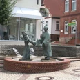 Brunnen - Die Wringerin Bronzefigur einer Frau beim Auswringen von Wäsche auf einem Brunnen in einem gepflasterten Stadtplatz.