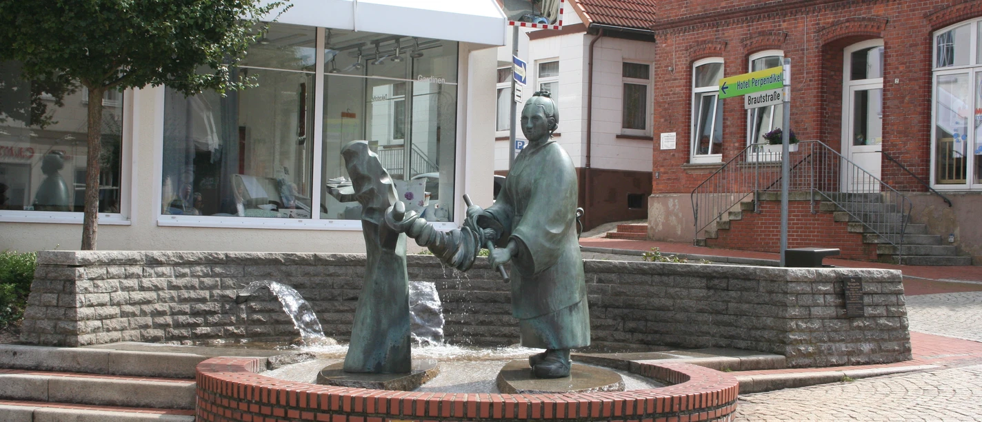Brunnen - Die Wringerin Bronzefigur einer Frau beim Auswringen von Wäsche auf einem Brunnen in einem gepflasterten Stadtplatz.