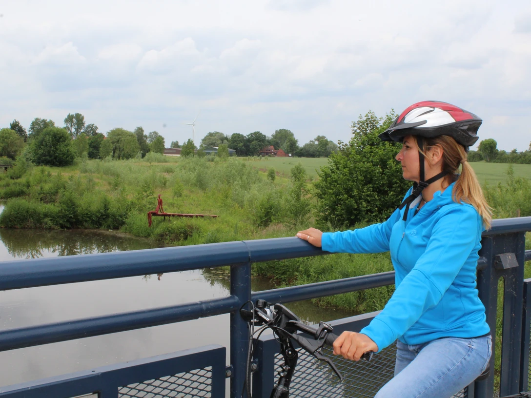 Radfahrerin mit Schutzhelm betrachtet von einer Brücke einen ruhigen Flusslauf inmitten grüner Natur.