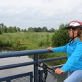 Radfahrerin mit Schutzhelm betrachtet von einer Brücke einen ruhigen Flusslauf inmitten grüner Natur.