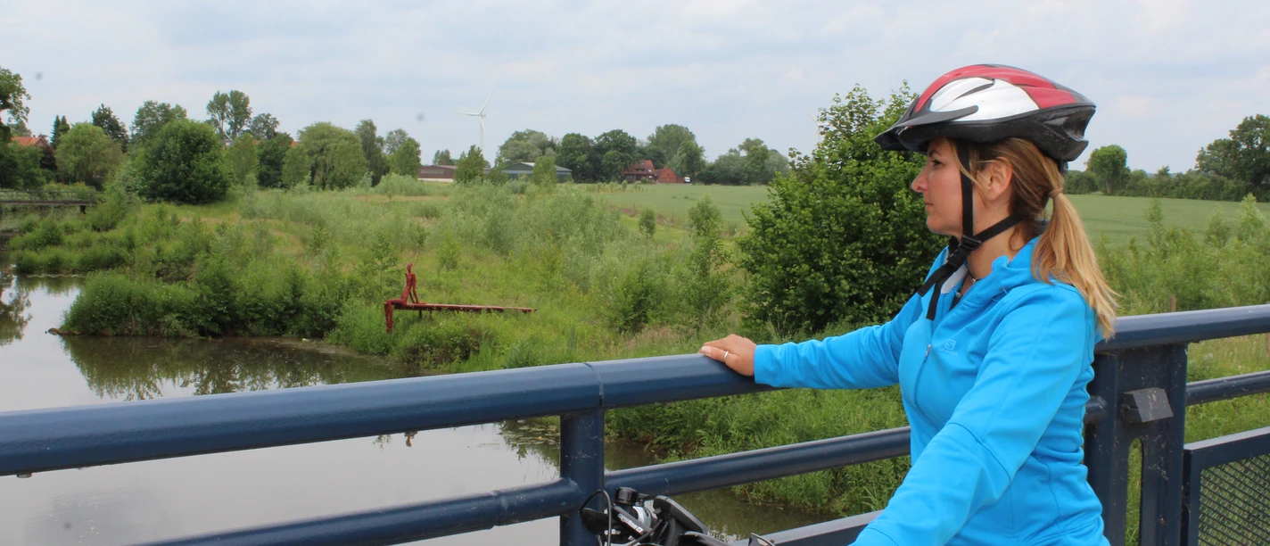 Radfahrerin mit Schutzhelm betrachtet von einer Brücke einen ruhigen Flusslauf inmitten grüner Natur.