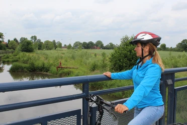 Meliorationstour Radfahrerin mit Schutzhelm betrachtet von einer Brücke einen ruhigen Flusslauf inmitten grüner Natur.