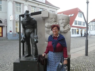 Eine Frau steht vor einer Skulpturengruppe mit einer knienden, arbeitenden Figur und lächelnden Babys.