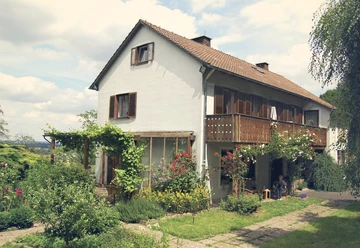 Bed und Breakfast Philosphenweg Haus aussen.jpg Ein zweistöckiges, weißes Haus mit Holzveranda, Garten und Sträuchern, eingebettet in eine grüne Landschaft.