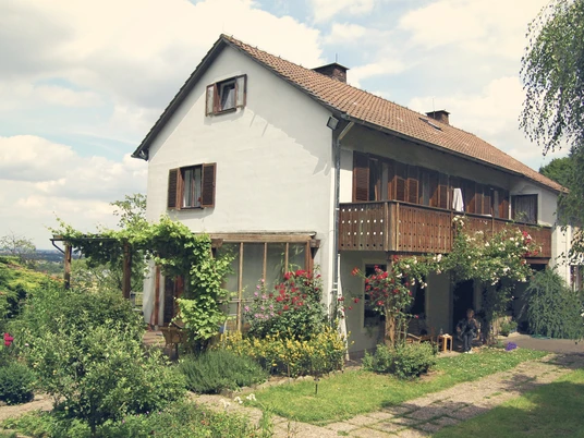 Bed und Breakfast Philosphenweg Haus aussen.jpg Ein zweistöckiges, weißes Haus mit Holzveranda, Garten und Sträuchern, eingebettet in eine grüne Landschaft.