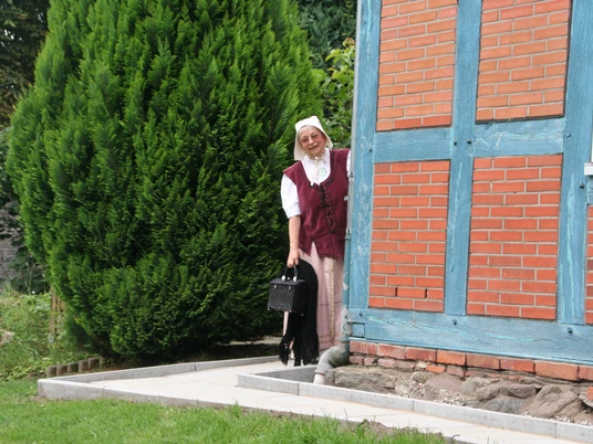 Eine Frau in historischer Tracht blickt lächelnd hinter einer Fachwerkfassade hervor, umgeben von Grün.